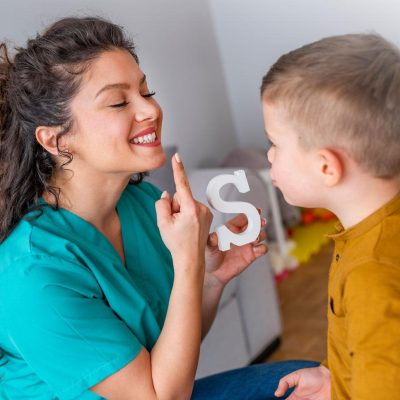 Female health care worker pointing at her smiling mouth while holding up the letter S in front of a young boy, teaching him how to make the sound for S.
