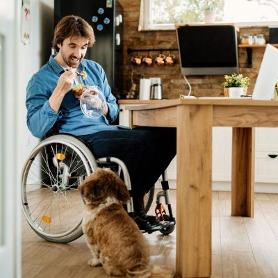 Man sitting in a wheel chair in front of a table, eating food while looking down at his small dog