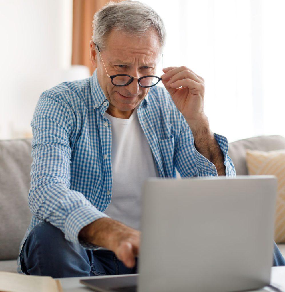 Senior man sitting on a couch looking at a computer while holding glasses up to his face.